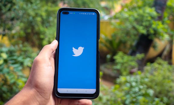 Hand holding a smartphone with the Twitter app open, outdoors with greenery in the background.