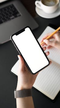 Hand holding a smartphone with a blank screen beside a notebook and laptop.